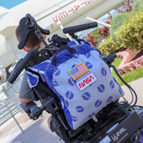 photo d'un fauteuil roulant de dos équipé d'un sac à dos décoré de patches, un enfant assis sur le fauteuil est partiellement visible de dos